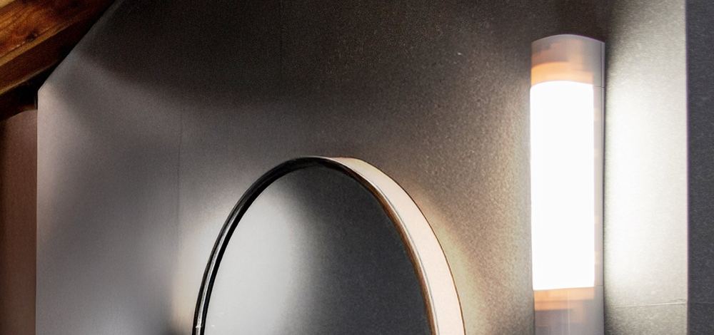 Réglette LED verticale allumée à côté d’un miroir rond dans une salle de bain
